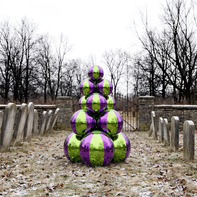 A stacked sculpture of reflective metallic spheres in alternating lime green and purple, placed outdoors in a grassy area with bare trees, stone walls, and gravestone markers.