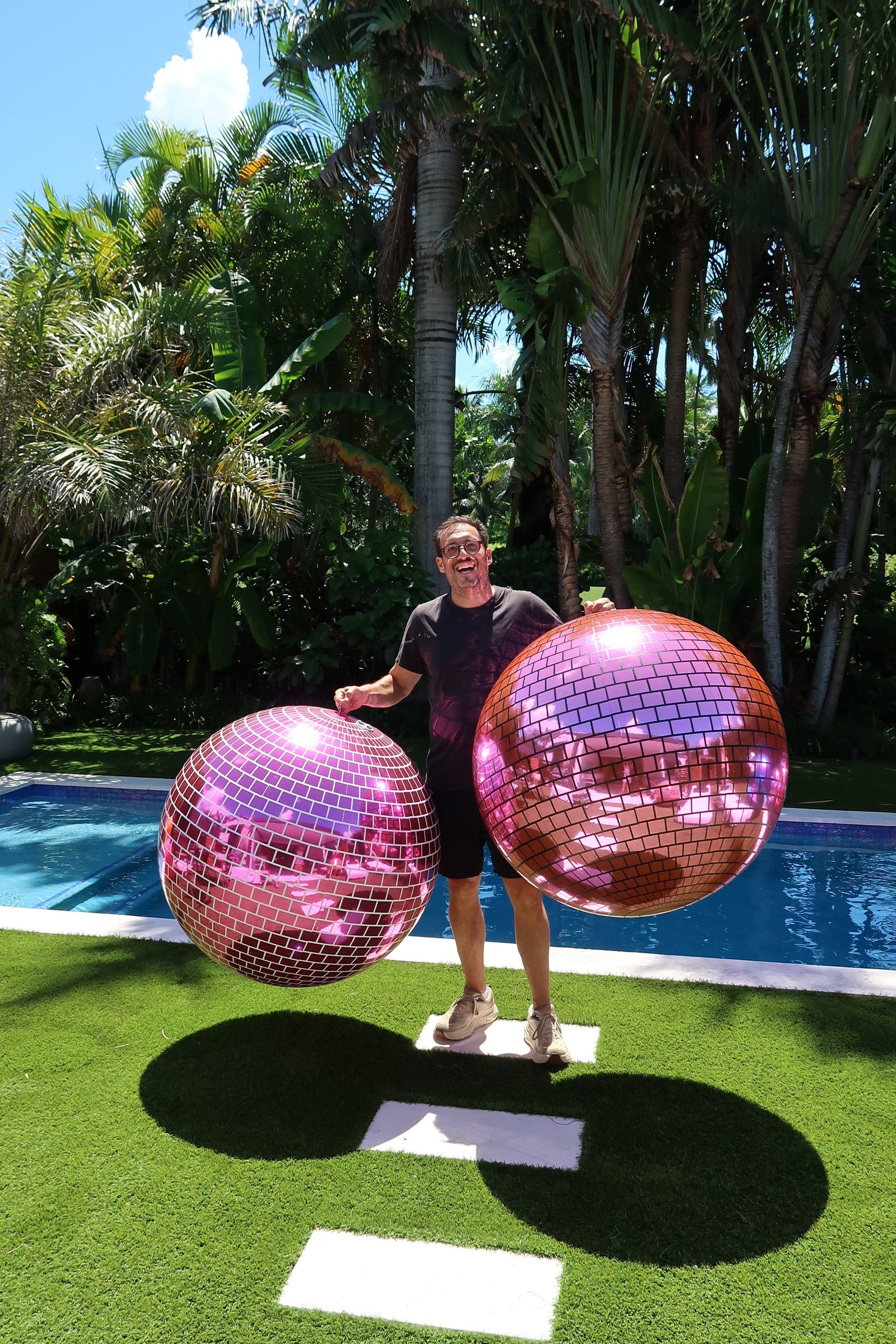 Person holding a large pink disco ball by a pool with palm trees in the background