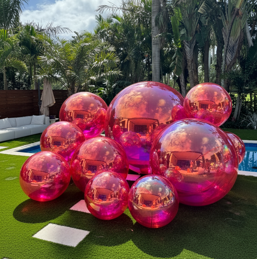 Pink and red transparent bubble spheres on a grassy area with palm trees and a pool in the background.