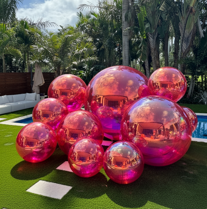 Pink and red transparent bubble spheres on a grassy area with palm trees and a pool in the background.