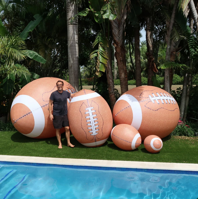 Man standing next to large inflatable footballs by a pool with palm trees in the background