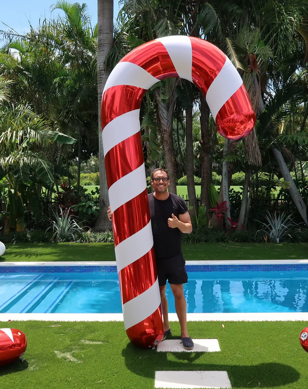 A person standing by a large inflatable shiny candy cane by a pool, with palm trees in the background.