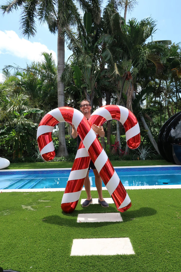 A person standing by a large inflatable shiny candy cane by a pool, with palm trees in the background.