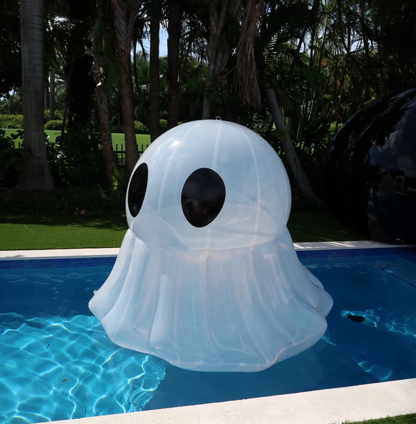 An inflatable ghost decoration floating in a pool, with a tropical setting in the background.