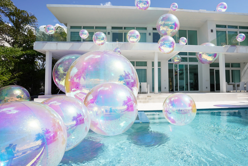 Inflatable chrome mirror-finish balls of various sizes arranged near a pool, with a modern house in the background.