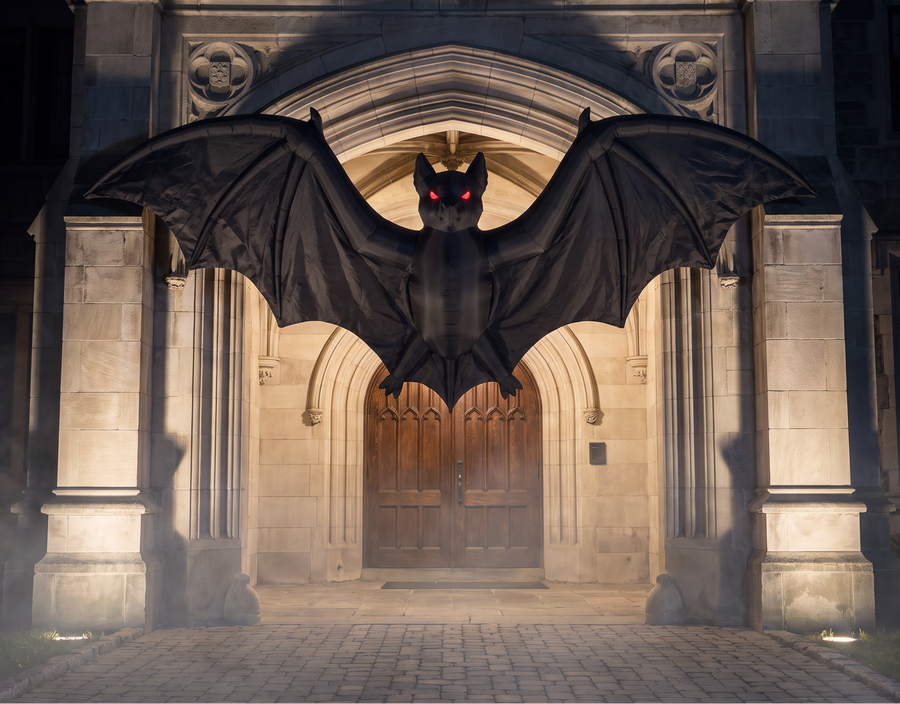 A large black bat decoration with red glowing eyes and a gold crown, suspended in front of a stone gothic building entrance at night.