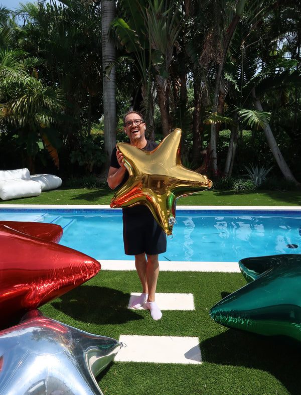 Person holding a gold star-shaped balloon by a pool with colorful balloons on the ground.