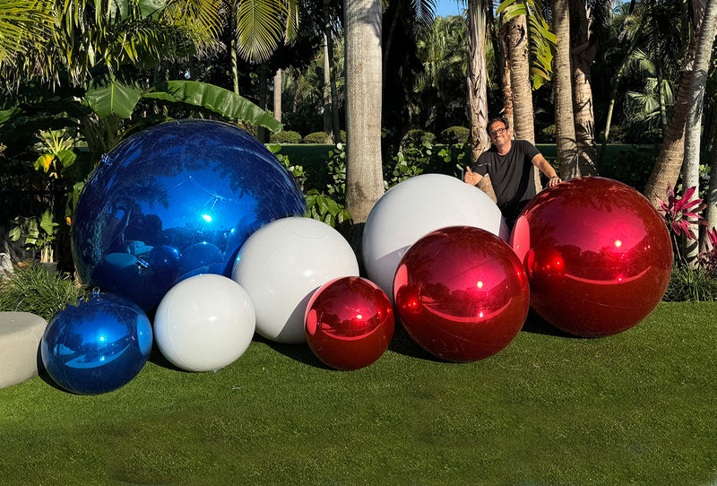 A collection of large metallic inflatable balls in red, white, and blue colors displayed outdoors with a person standing behind them.