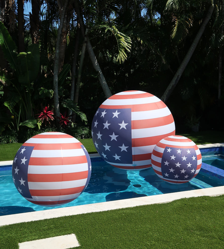 Three inflatable American flag balls by a pool with greenery in the background