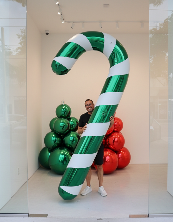 Person standing next to a large inflatable candy cane and Christmas tree balloons in a store window.